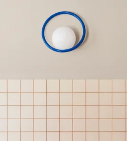 Luminaire type applique de la marque SWEDISH NINJA, en décoration sur un mur de teinte pèche pâle et une faïence décorative. Le luminaire est une boule sur laquelle est fixée un anneau bleu électrique qui tranche avec le fond