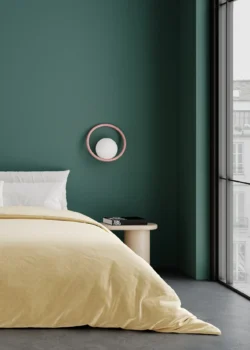 Luminaire type applique de la marque SWEDISH NINJA qui est mis en décoration dans une chambre composée d'un mur peint en vert et d'un lit. Le luminaire est une boule surmontée d'un année de teinte rose pale qui tranche avec le fond