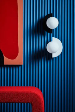 Deux luminaires type applique de la marque SWEDISH NINJA sont accrochés de manière décorative, sur un mur bleu électrique texturé. On trouve aussi des éléments de décoration rouge vif. Les deux luminaires sont des boules agrémentées de demis cercles métalliques de teinte blanche et noire